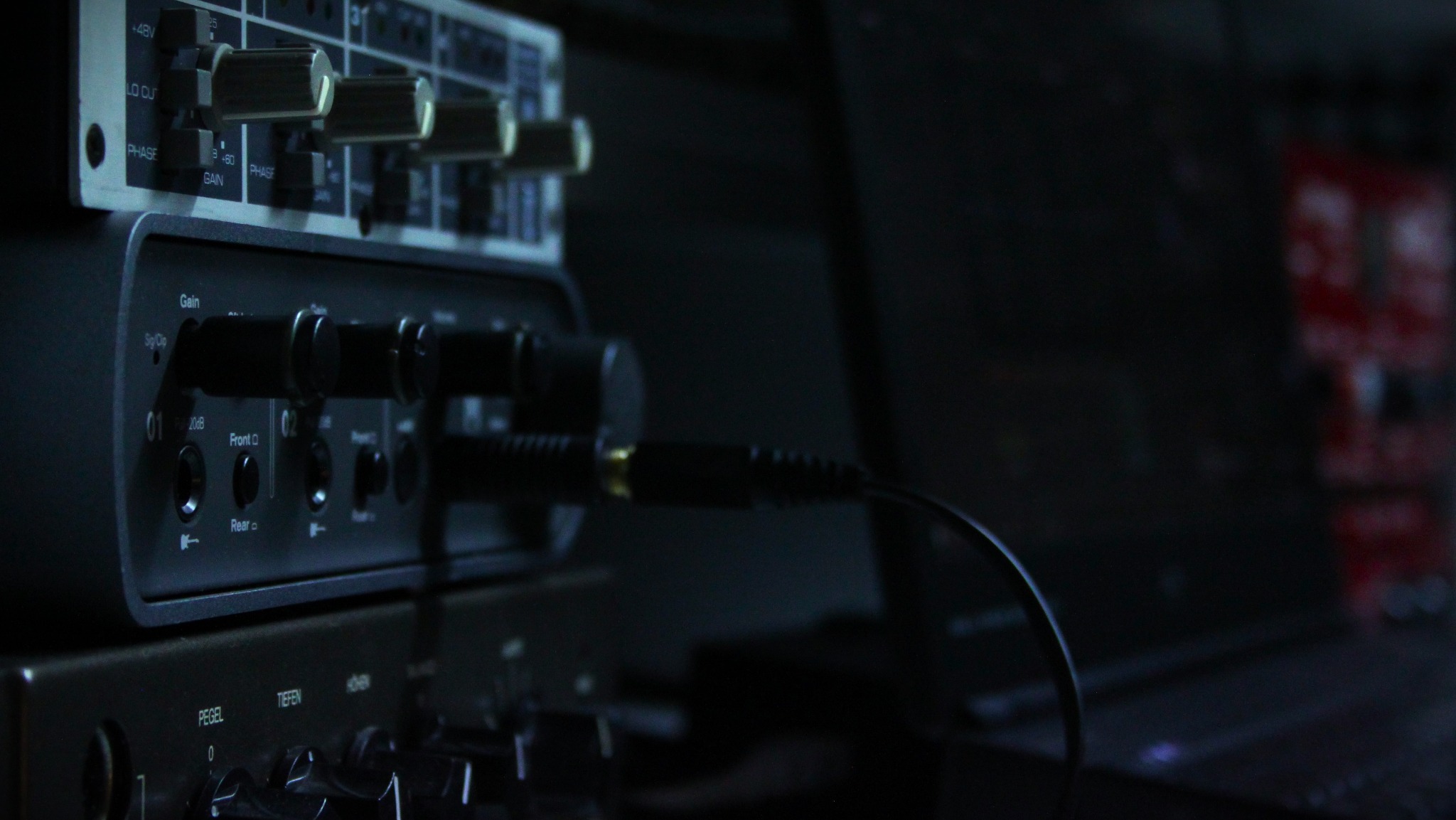 Sprzęt studyjny: interfejs audio z wpiętymi kablami, mikser i laptop w tle. Elementy sterujące, potencjometry i gniazda widoczne w zbliżeniu. Nastrojowe, ciemne oświetlenie.