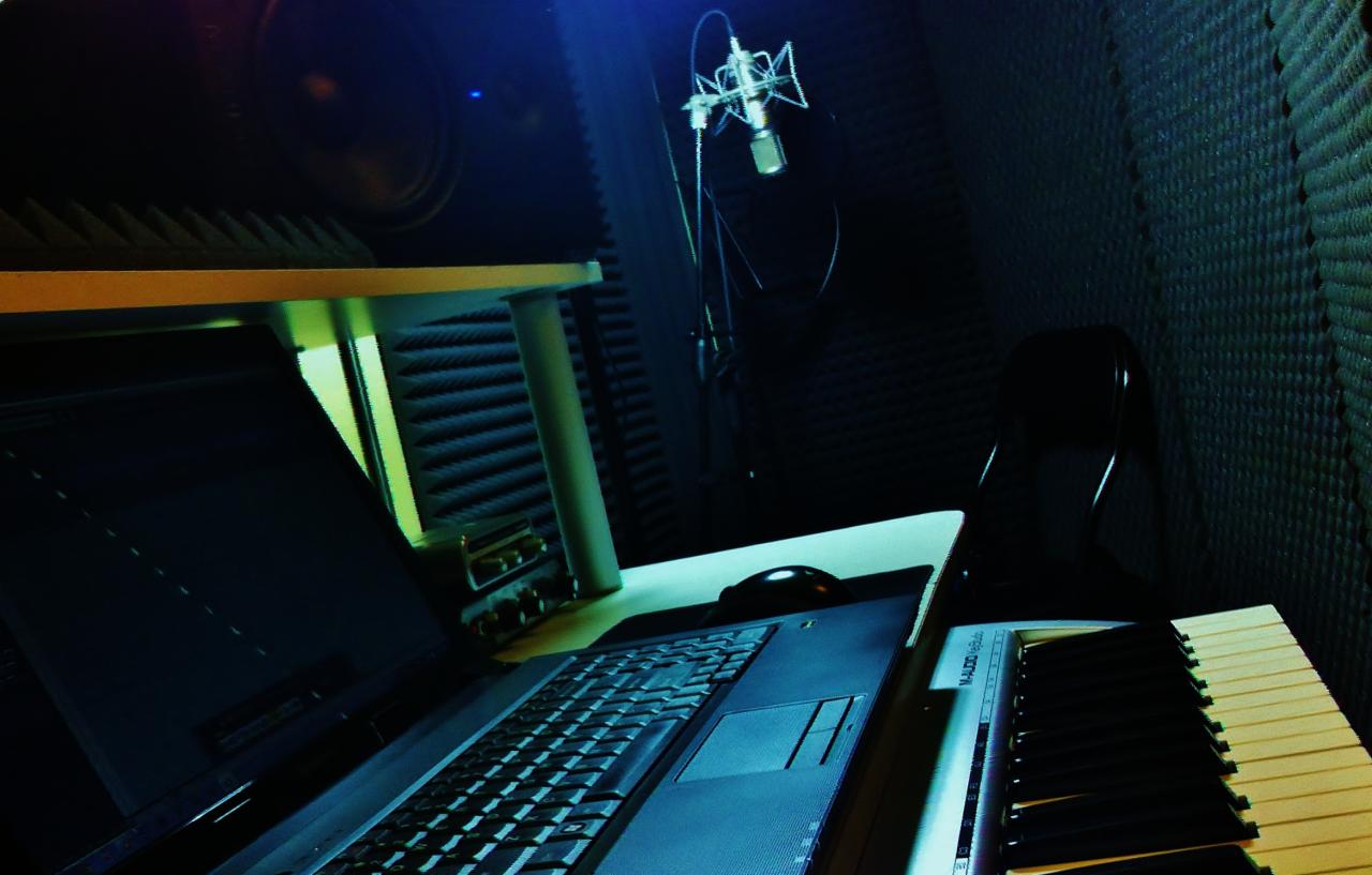 Wnętrze domowego studia nagrań z mikrofonem, laptopem i klawiaturą MIDI. Ściany wyłożone panelami akustycznymi. Nastrojowe oświetlenie.
