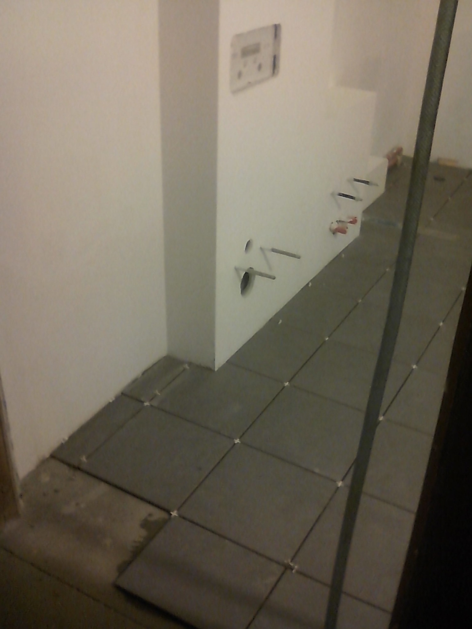 Układanie szarych płytek podłogowych w narożniku pomieszczenia, widoczne rury wystające ze ściany i krzyżyki dystansowe między płytkami.