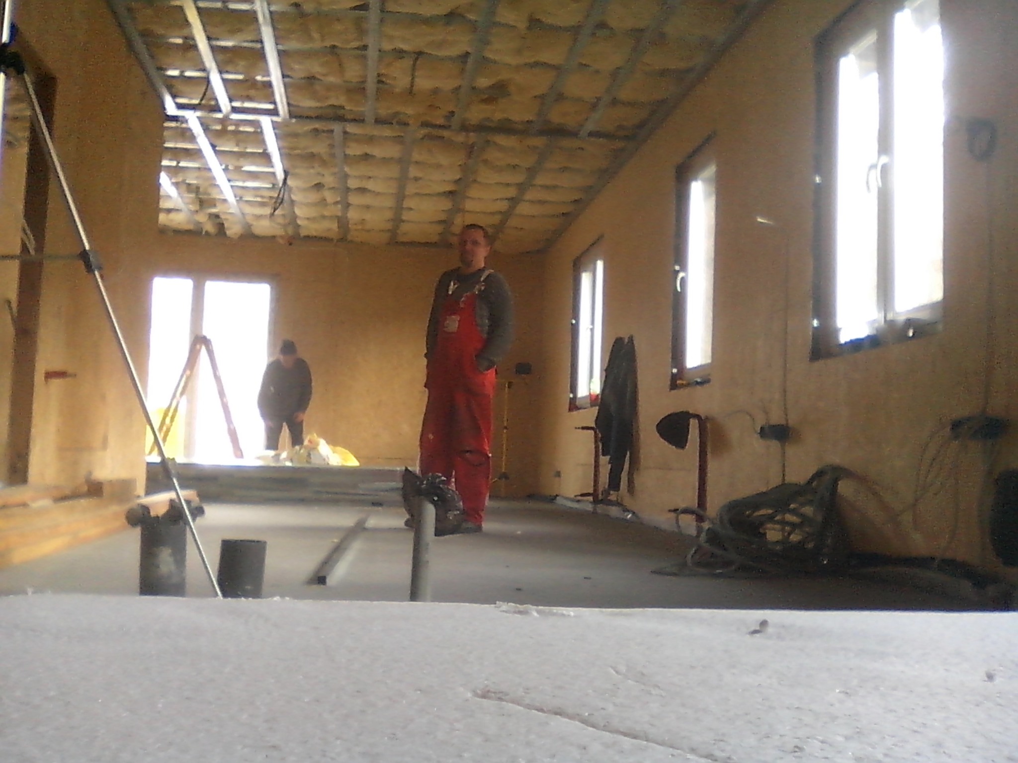 Wnętrze w trakcie prac: montaż izolacji poddasza z widoczną konstrukcją rusztu, pracownik w czerwonym kombinezonie i materiał izolacyjny na pierwszym planie.