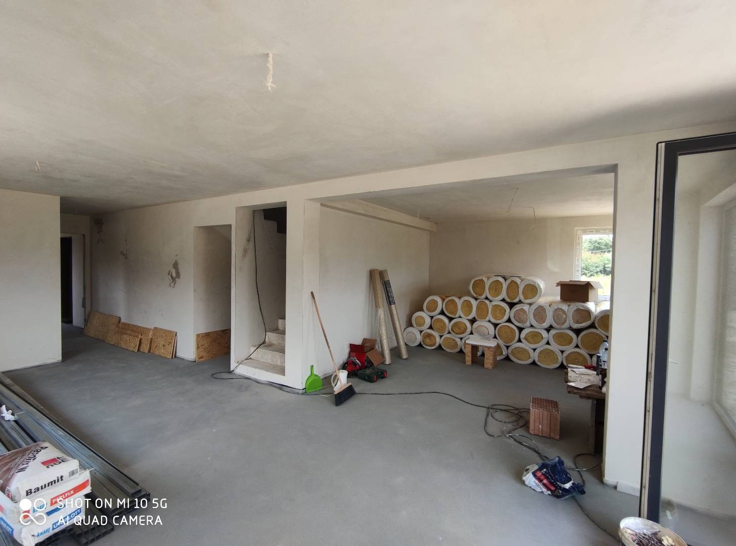 Wnętrze w trakcie prac przygotowawczych: surowe ściany, betonowa podłoga, materiały izolacyjne w rolkach zgromadzone w rogu pomieszczenia, widoczne narzędzia i akcesoria budowlane.