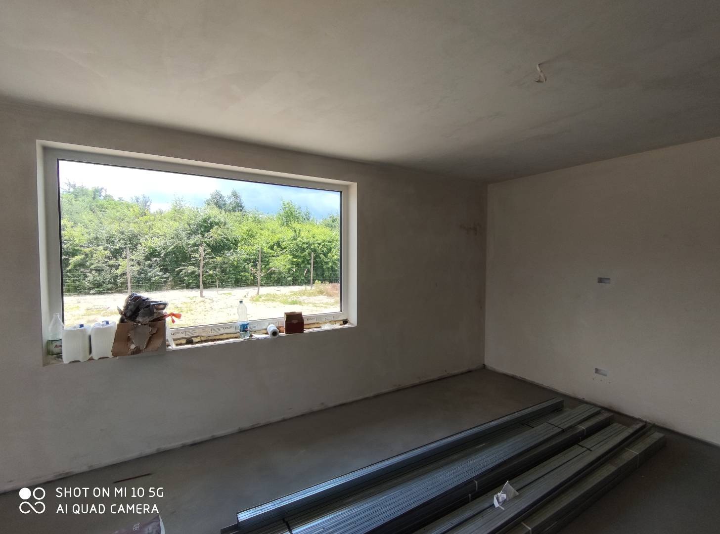 Wnętrze w stanie surowym po tynkowaniu maszynowym, widoczne profile metalowe na podłodze i zaplecze materiałów przy oknie, ściany i sufit pokryte świeżym tynkiem.