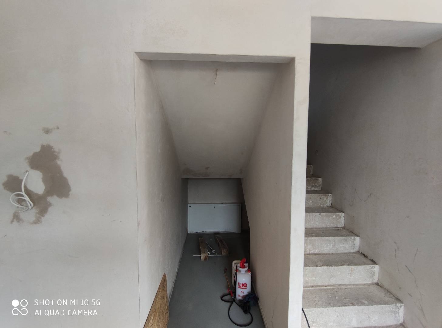 Surowe wnętrze w trakcie prac wykończeniowych: ściany po nałożeniu warstwy wyrównującej, nieotynkowane schody, widoczny spryskiwacz i kabel wystający ze ściany.