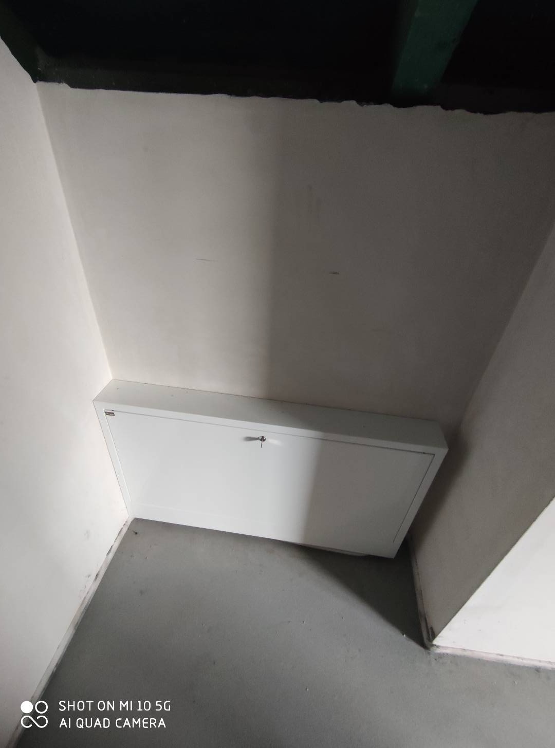 Narożne ujęcie białej szafki rewizyjnej z kluczykiem, zamontowanej w ścianie, widoczna szara podłoga i fragment sufitu z elementami instalacji.