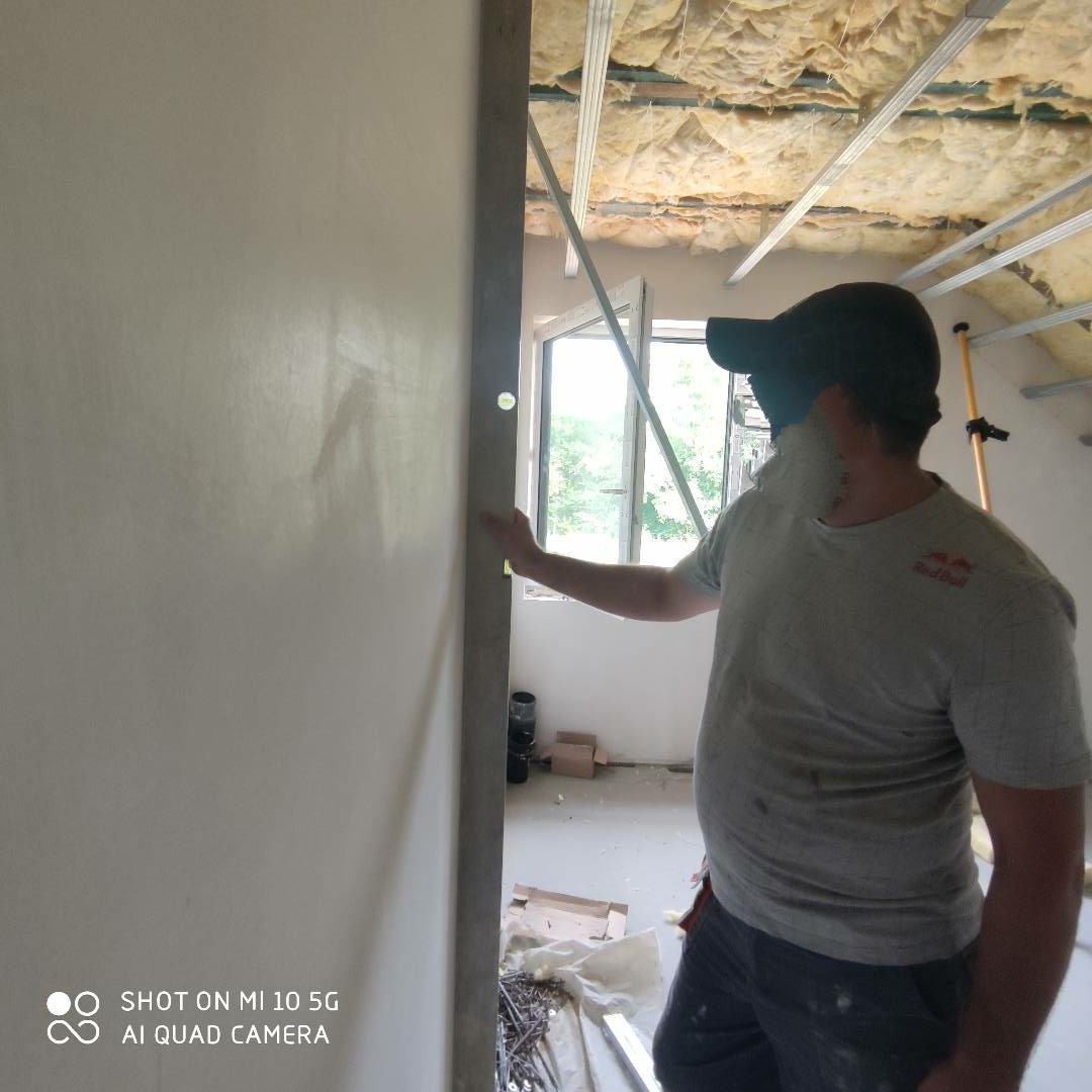 Pracownik w czapce sprawdza pion pionową poziomicą na świeżo wykończonej, jasnej ścianie. Widoczne ocieplenie poddasza i okno w tle.