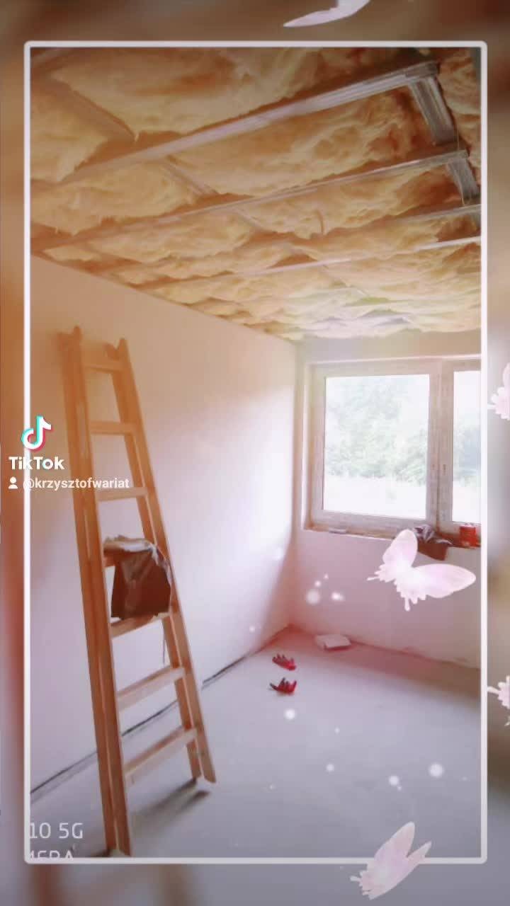 Poddasze w trakcie ocieplania, widoczna drabina oparta o ścianę, częściowo zamontowana izolacja w metalowym stelażu podwieszanego sufitu, okno w tle.