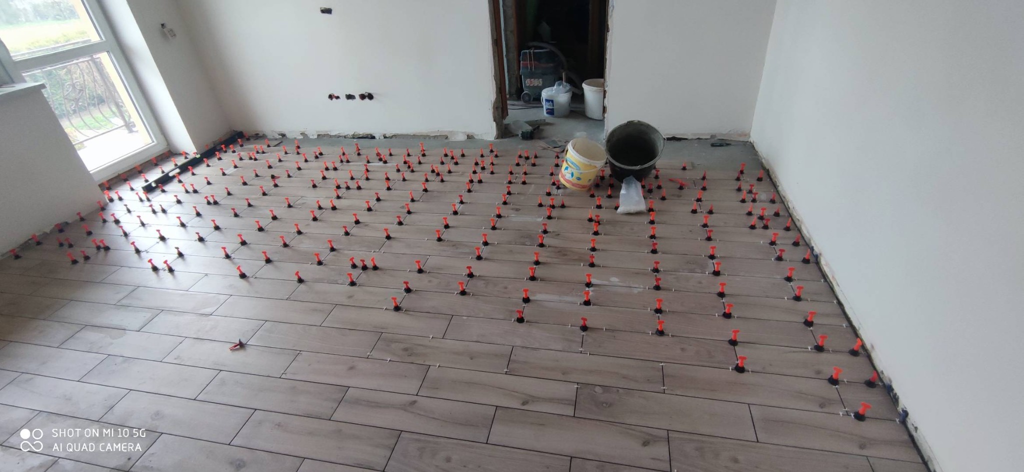 Układanie płytek podłogowych z użyciem systemu poziomowania, widoczne pomarańczowe kliny i białe krzyżyki dystansowe, w tle dwa wiadra i worki z materiałami budowlanymi.