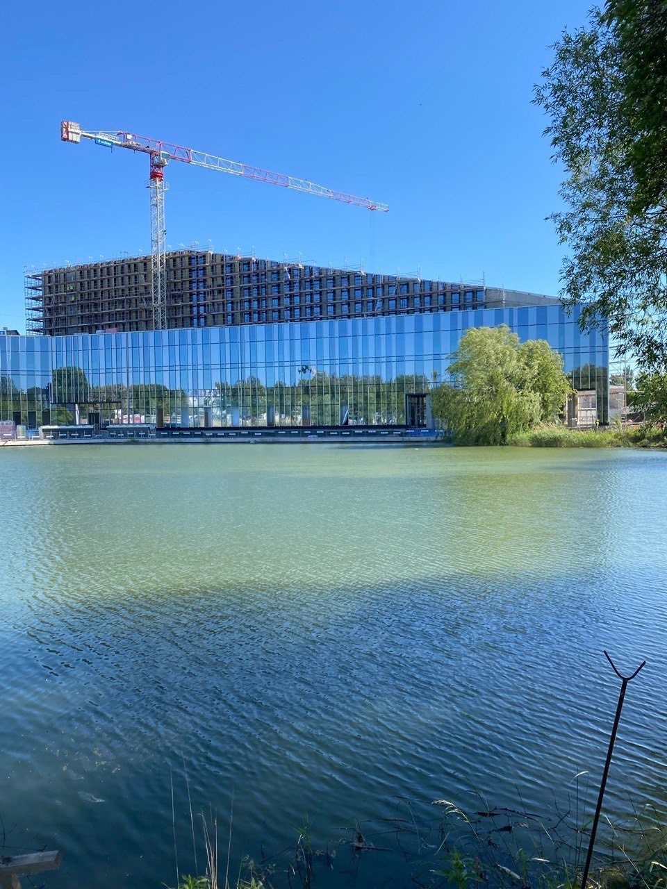 Budynek żelbetowy w trakcie budowy, widoczny żuraw budowlany, odbicie w tafli wody. Nowoczesna architektura z elementami szkła.