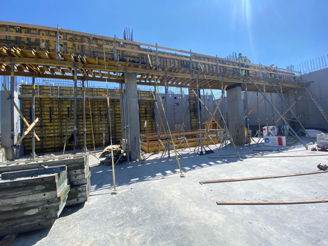 Żółte szalunki Doka na budowie, wsparte na podporach, z widocznymi zbrojeniami i pracownikami na górze, w słoneczny dzień. Elementy konstrukcji betonowej.