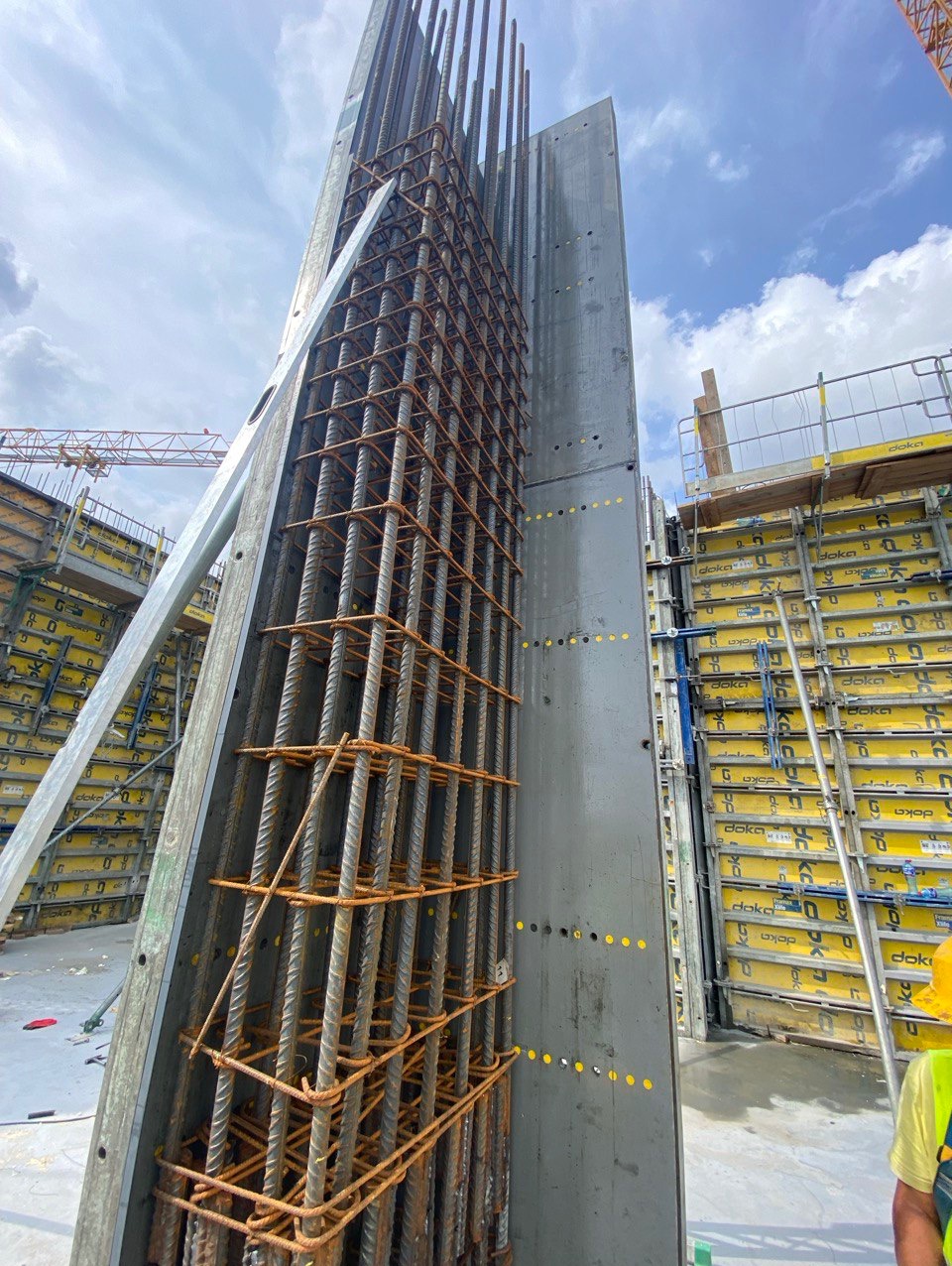 Zbrojenie pionowe w szalunku, widok z dołu na konstrukcję z prętów stalowych. Szalunek z otworami, w tle niebo i inne elementy konstrukcji.