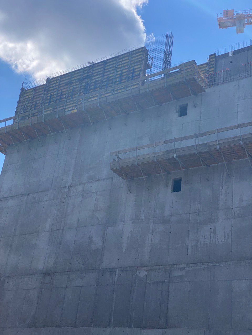 Wysoki budynek żelbetowy w trakcie budowy, widoczne szalunki i zbrojenia na tle błękitnego nieba z chmurami. Na górze kadru widoczny żuraw budowlany.