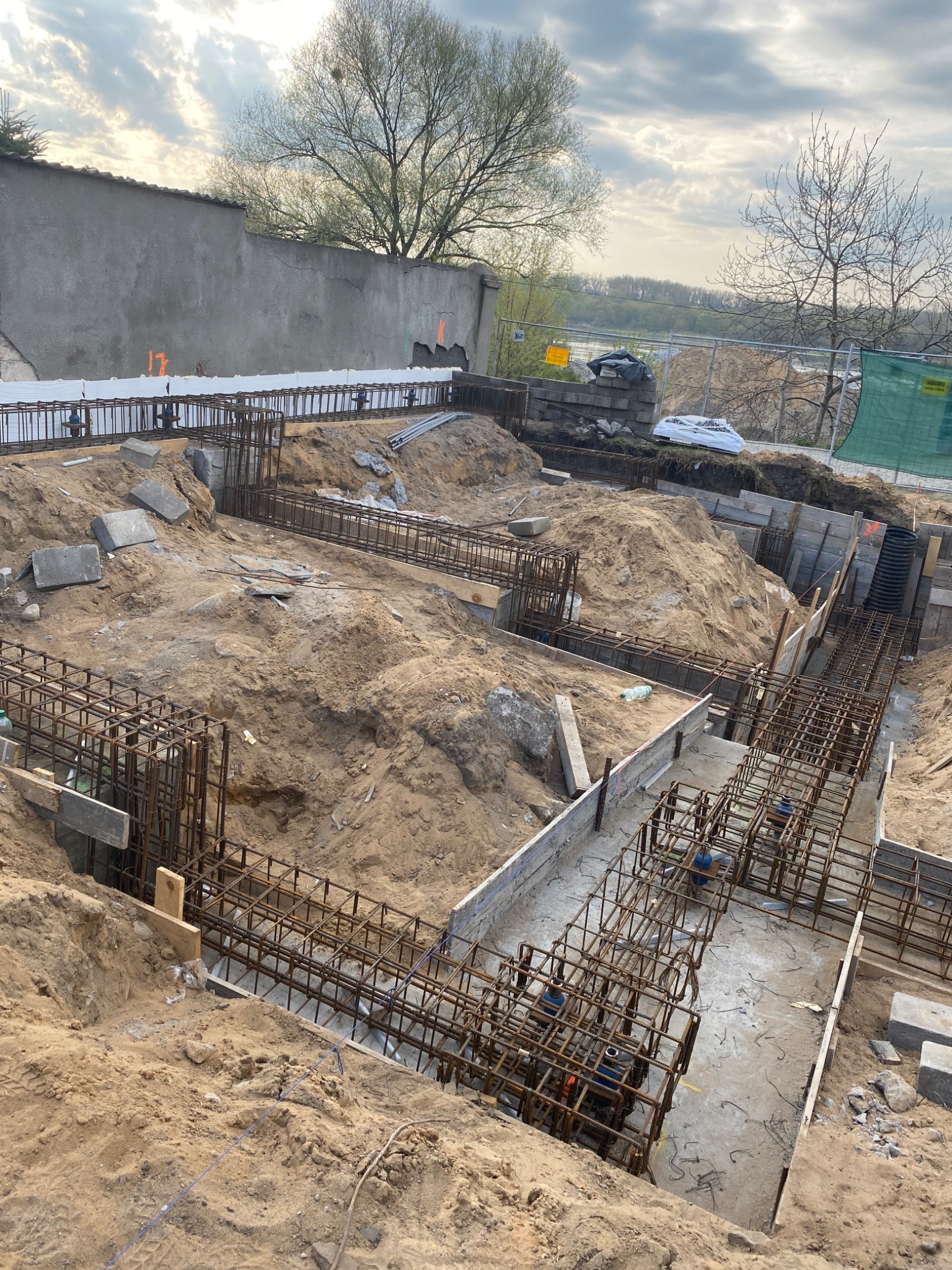 Szeroki kadr wykopu z przygotowanym zbrojeniem i szalunkiem pod fundamenty budynku jednorodzinnego, widoczne wystające pręty zbrojeniowe, elementy izolacji termicznej oraz ziemia.