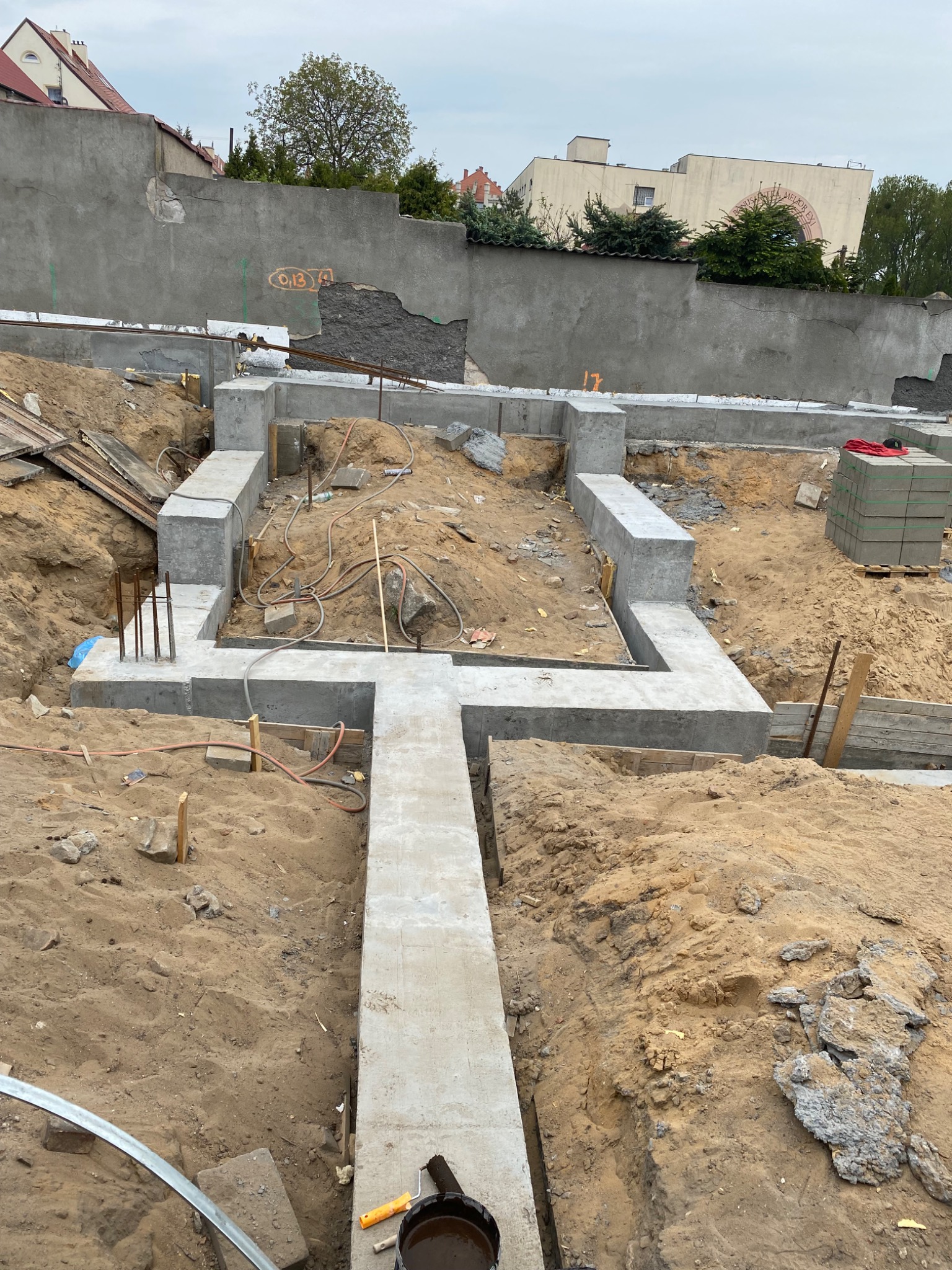 Wykop pod fundamenty domu jednorodzinnego z wylanym betonem, widoczne wystające pręty zbrojeniowe, przewody i narzędzia budowlane na piaszczystym podłożu, w tle fragment muru z oznaczeniem wysokości.