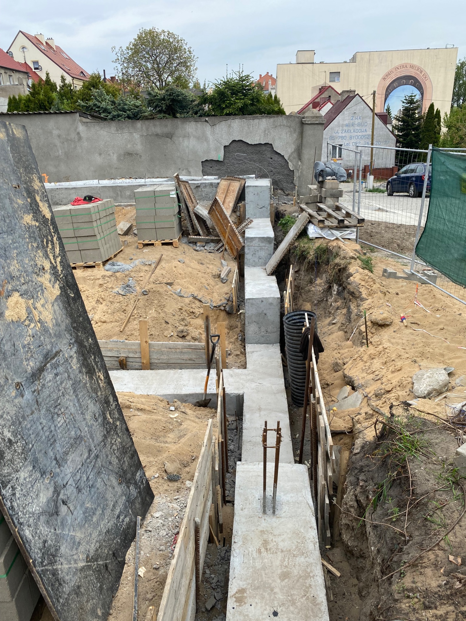 Wykop pod ławy fundamentowe z częściowo wylanym betonem, widoczne szalunki, zbrojenie oraz rury kanalizacyjne w wykopie, w tle mur z ubytkami tynku i budynki mieszkalne.