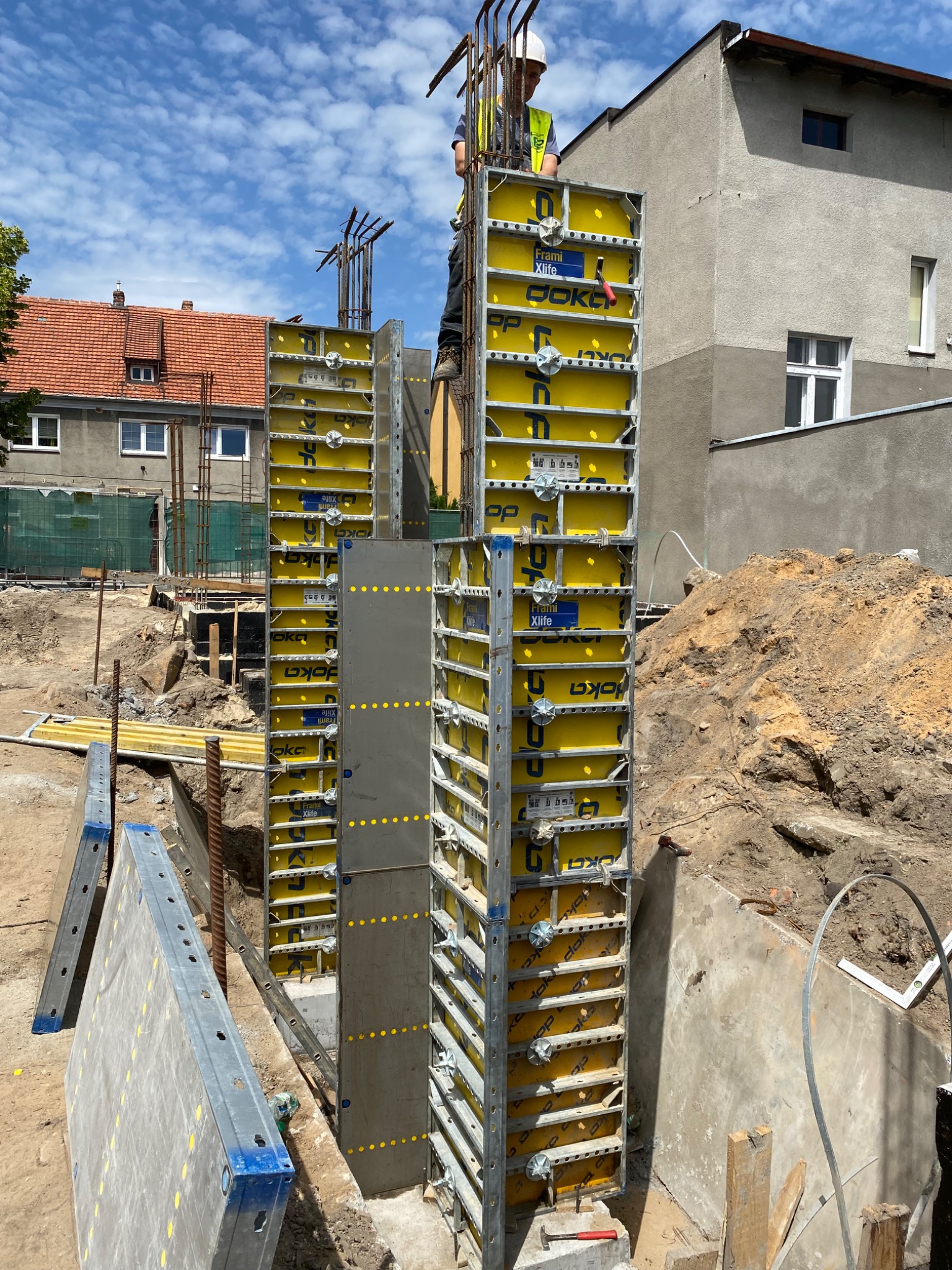 Budowa słupów żelbetowych z użyciem żółtych szalunków systemowych Doka, widoczny pracownik w kasku i kamizelce odblaskowej na górze konstrukcji, zbrojenie wystające ponad szalunki, w tle budynki...