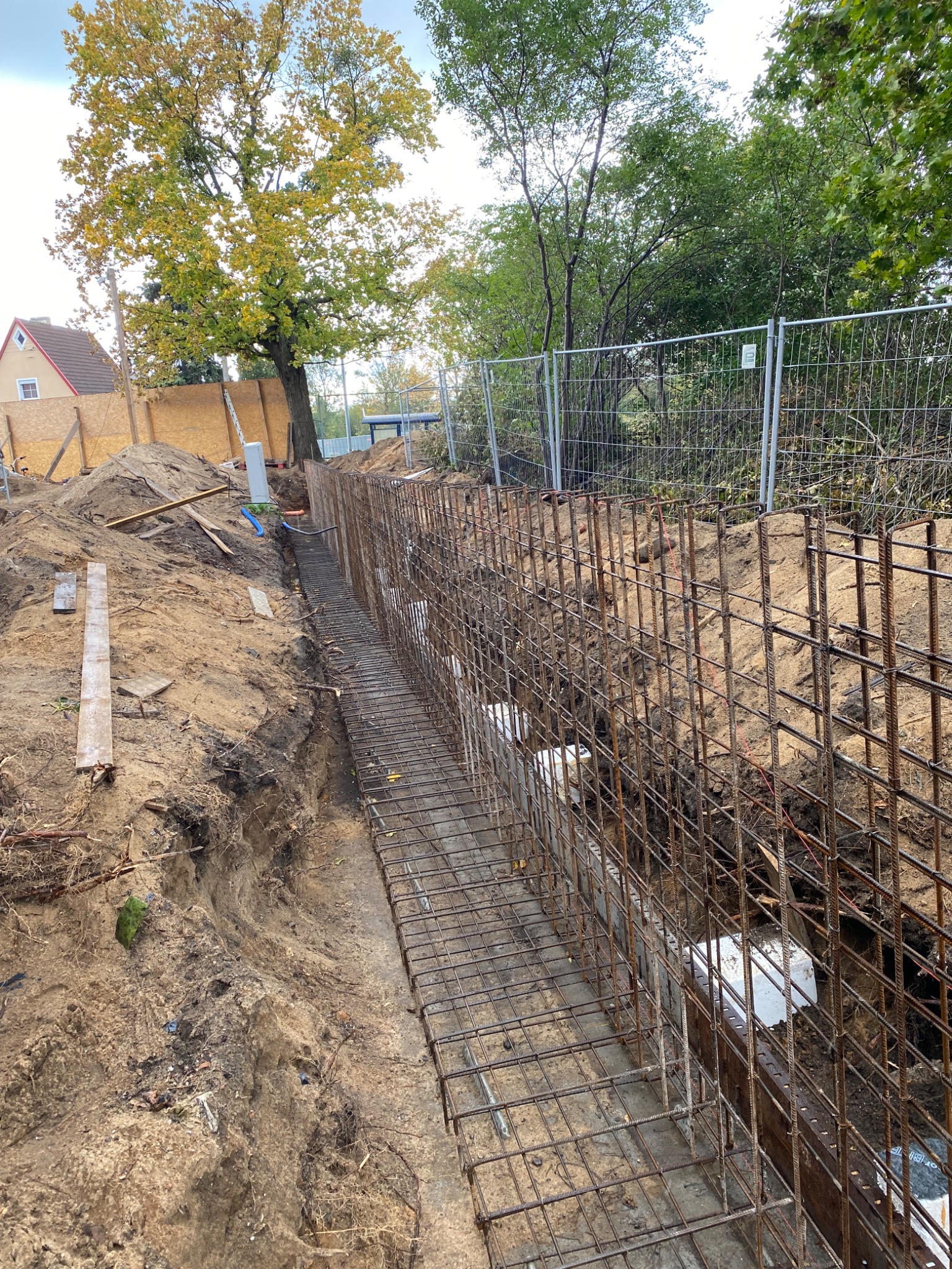 Wykop pod fundament z ułożonym zbrojeniem w formie siatki stalowej, widoczne elementy szalunku, w tle ogrodzenie budowy i drzewa.