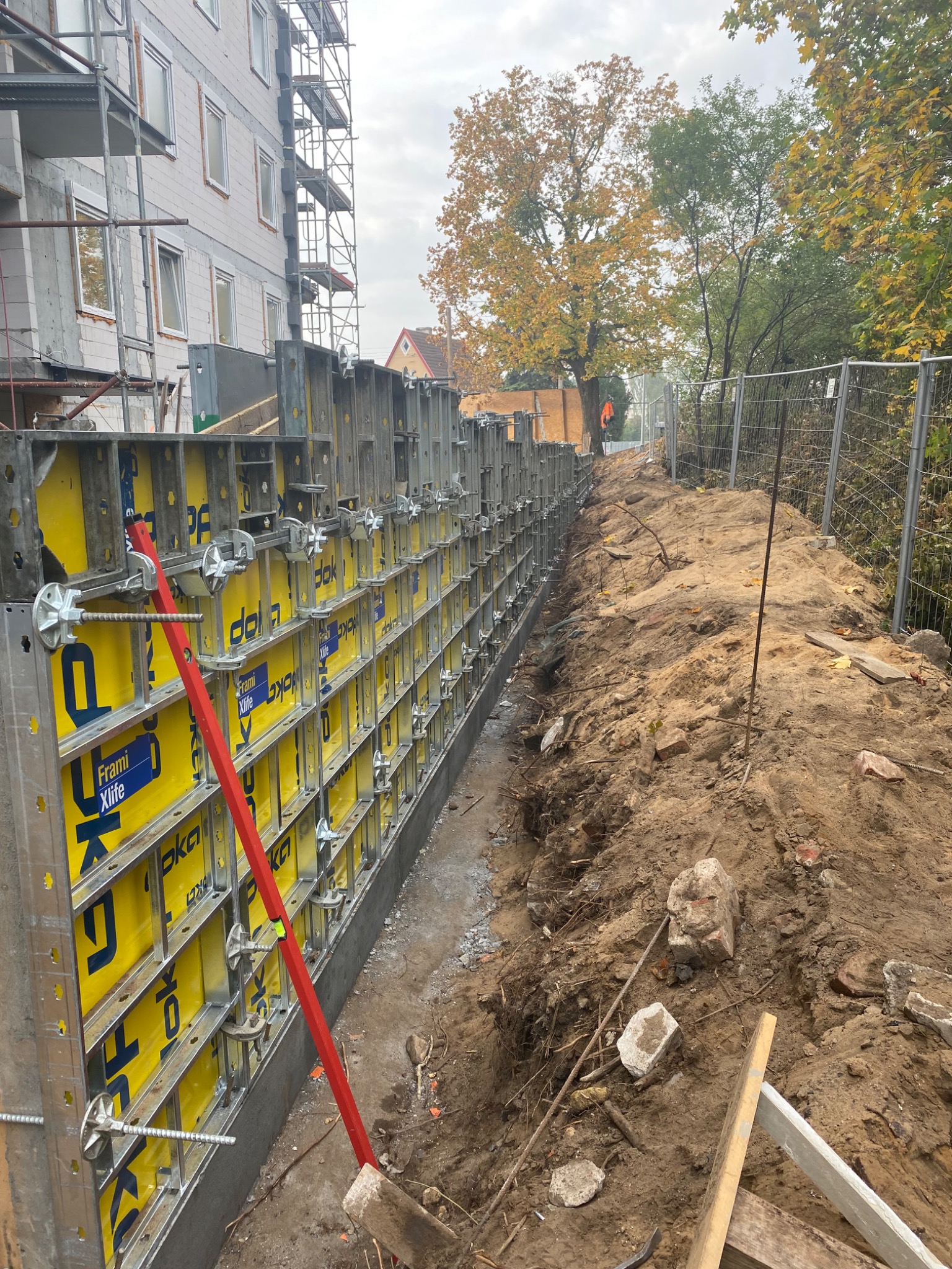 Szczegółowe ujęcie szalunku z żółtymi panelami i metalową konstrukcją, ustawionego w wykopie obok budynku w trakcie budowy. Czerwona poziomica oparta o szalunek. W tle widoczny grunt i drzewa.