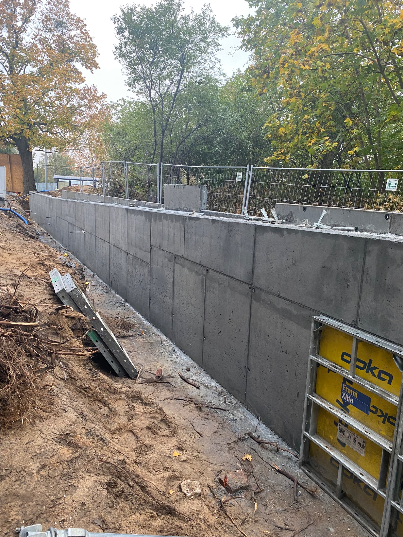 Świeżo wylany, segmentowy fundament betonowy z widocznymi elementami szalunkowymi i metalowymi akcesoriami mocującymi, w tle siatka zabezpieczająca i drzewa z jesiennymi liśćmi.