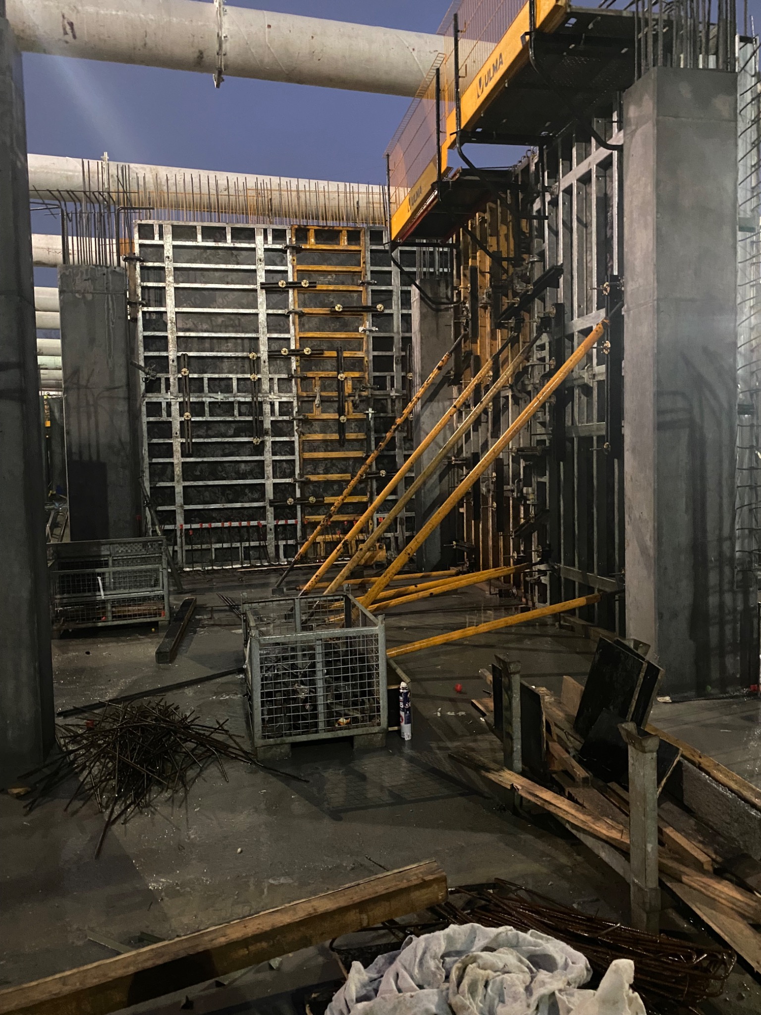 Budowa filaru z szalunkiem i zbrojeniem, widoczne elementy konstrukcyjne ULMA, pręty zbrojeniowe i materiały budowlane na placu budowy. Nocne ujęcie.