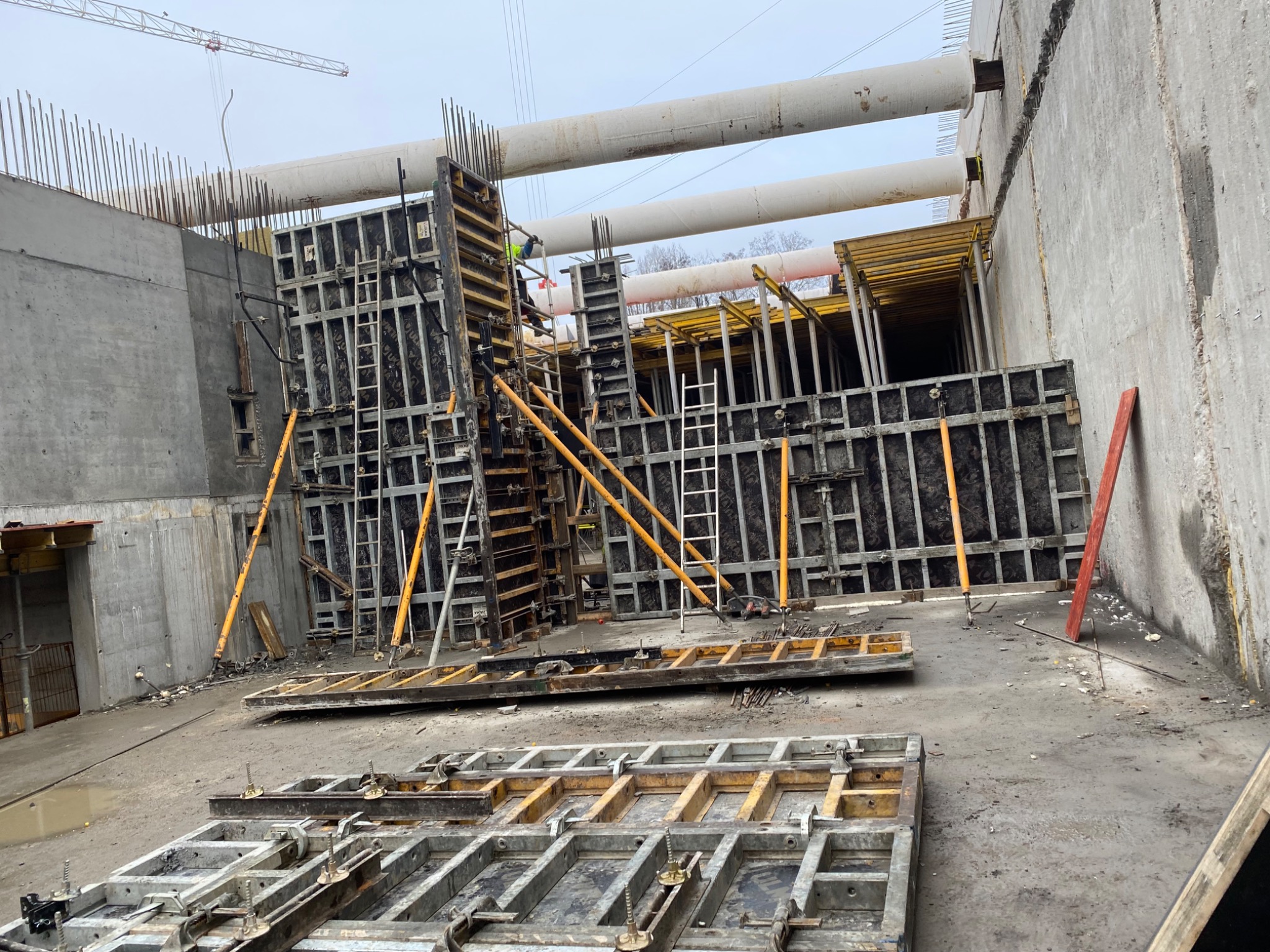 Szare betonowe ściany wykopu z widocznymi szalunkami, metalowymi elementami konstrukcyjnymi i rurami na górze, przygotowane do wylewania betonu.