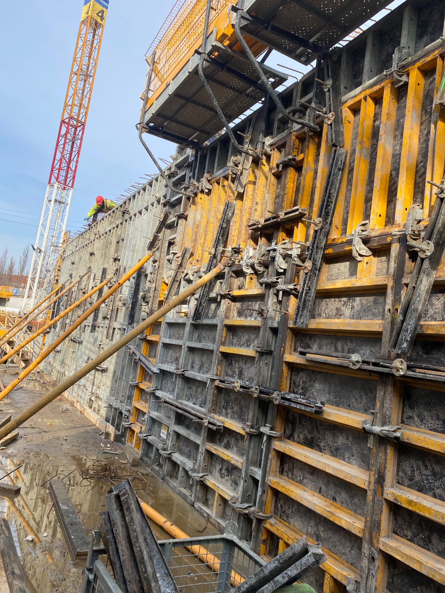 Żółte szalunki pionowe z metalowymi zaciskami na budowie, widoczny pracownik w czerwonym kasku na górze konstrukcji i żuraw w tle.