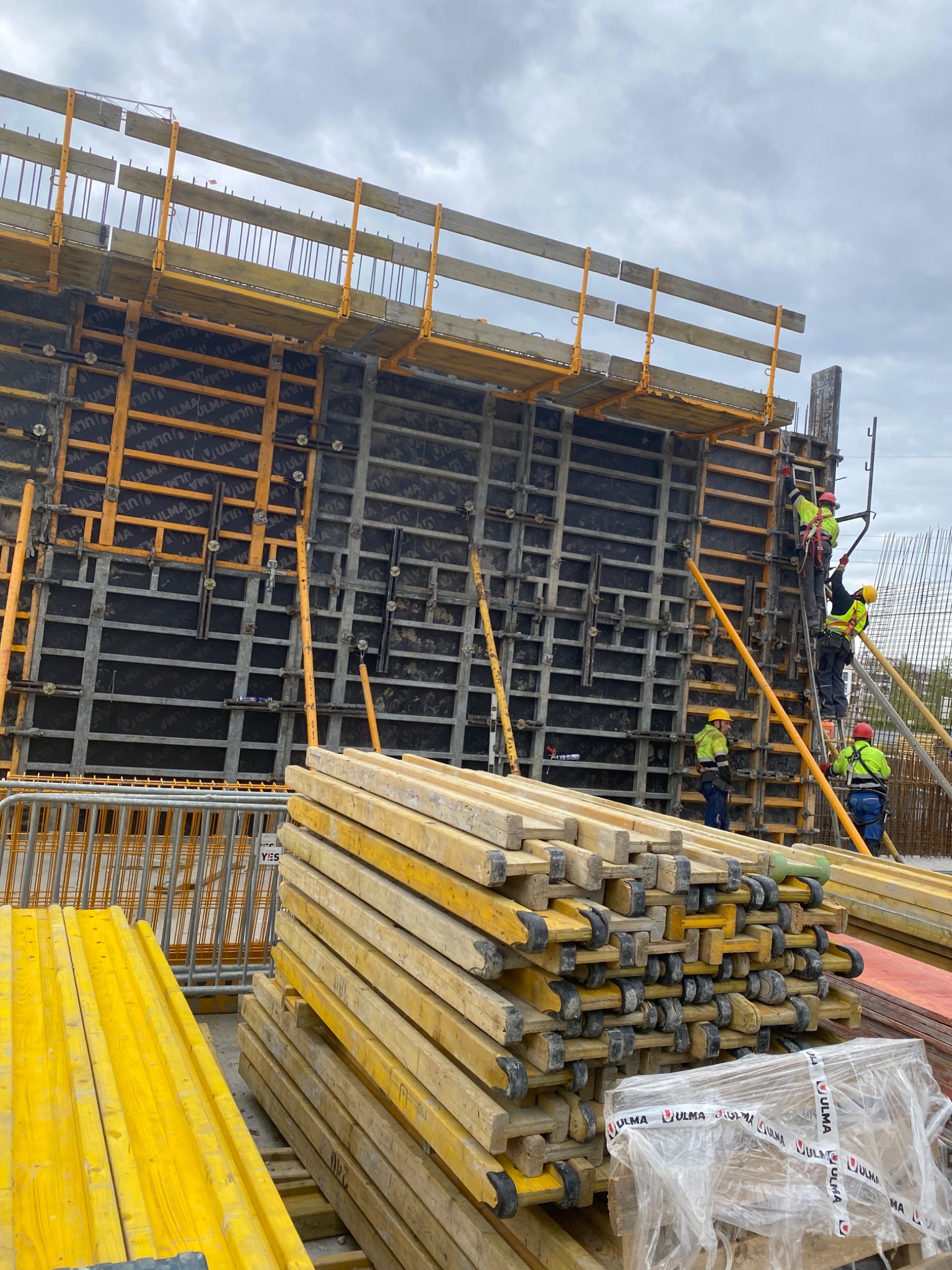 Prace budowlane: montaż szalunku na budowie z widocznymi pracownikami w odblaskowych kamizelkach, stosami drewnianych belek szalunkowych i zbrojeniem w tle.