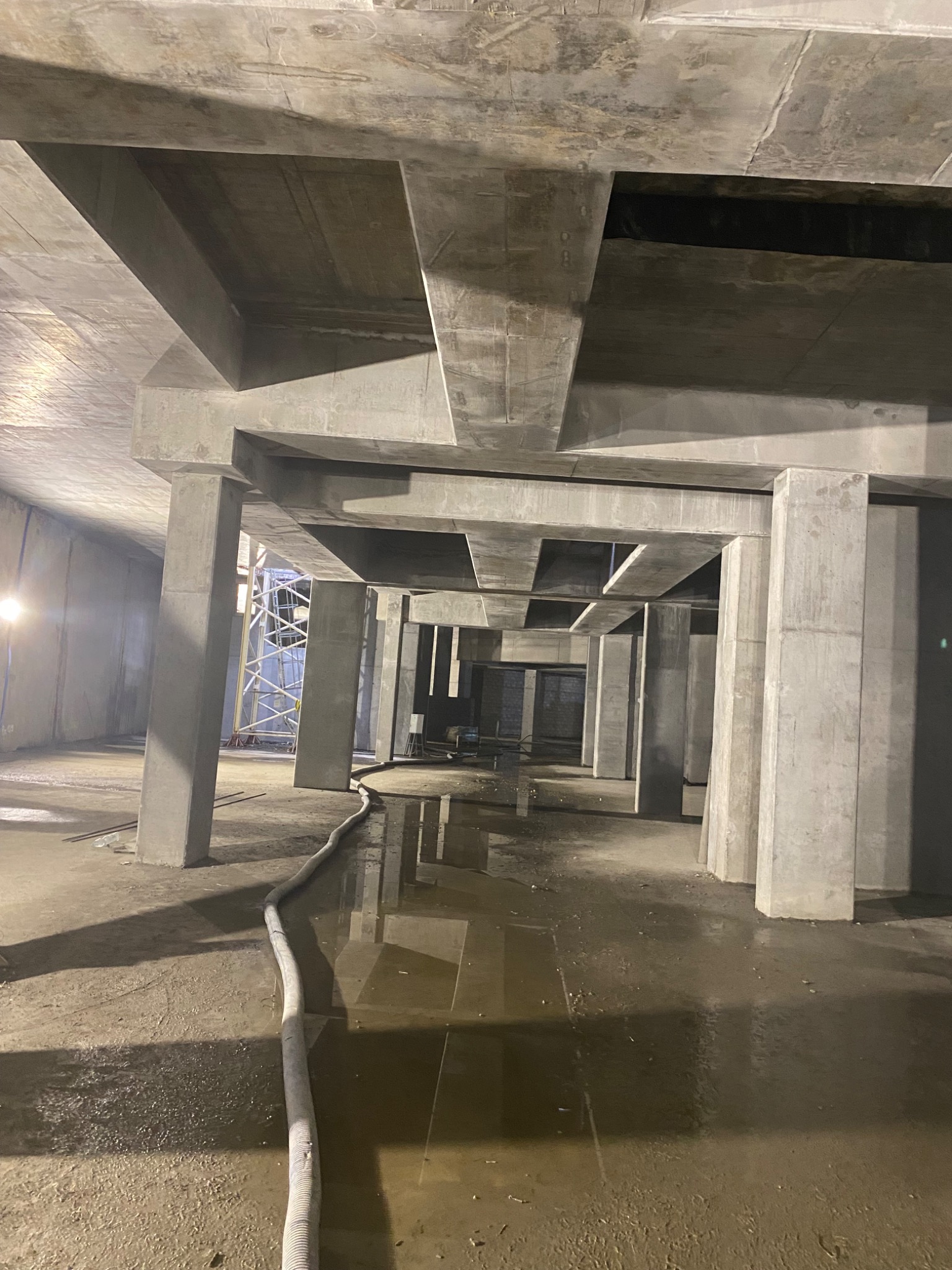 Wnętrze surowego budynku żelbetowego w trakcie budowy, widoczne liczne betonowe kolumny i strop z charakterystycznymi żebrami, na podłodze woda i rozłożony wąż, w tle rusztowanie.