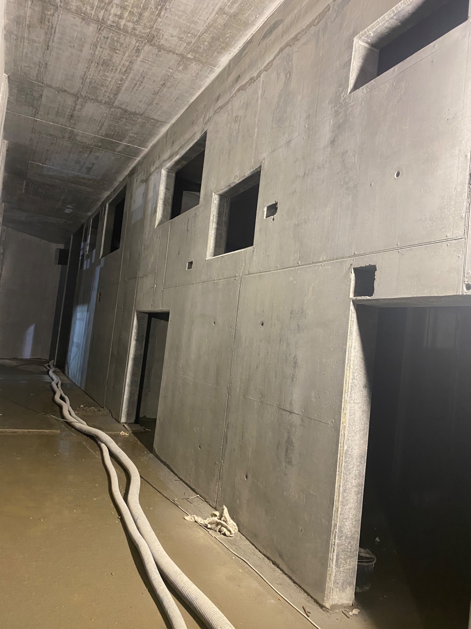 Surowy, betonowy szkielet budynku z otworami na okna i drzwi, widoczne szare rury na mokrej posadzce i niedokończone wykończenie sufitu.