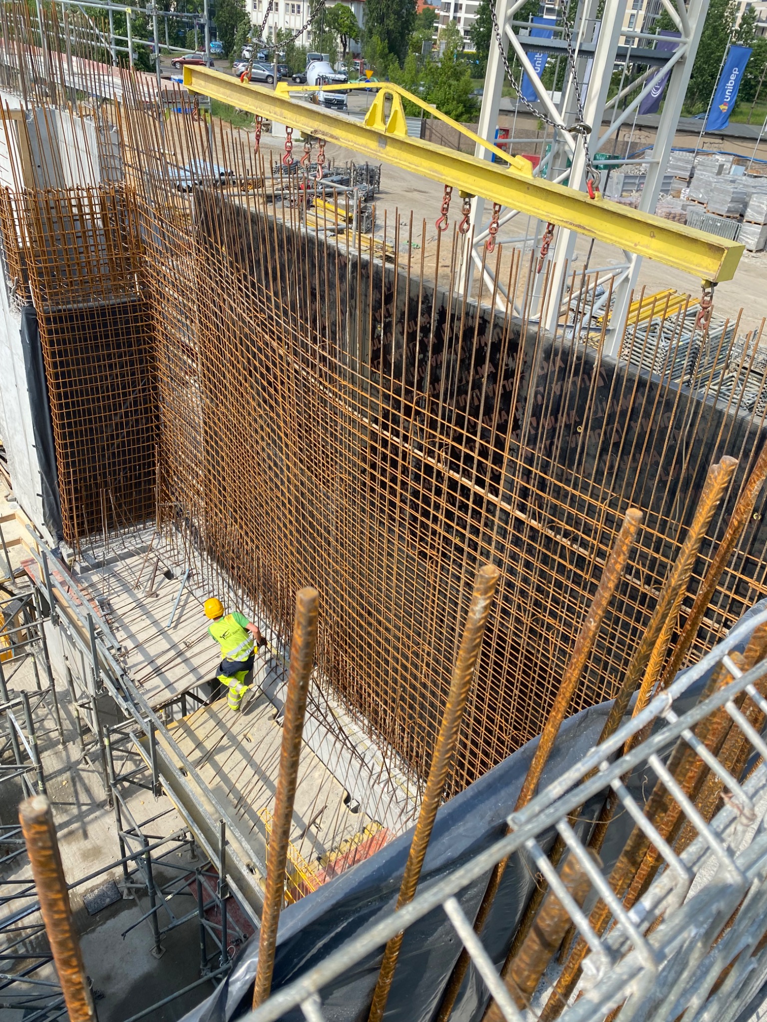 Widok z góry na plac budowy: pracownik w żółtym kasku i kamizelce na rusztowaniu, obok pionowa ściana z gęstej siatki zbrojeniowej, podnoszona przez żółty dźwig, w tle budynki miejskie.