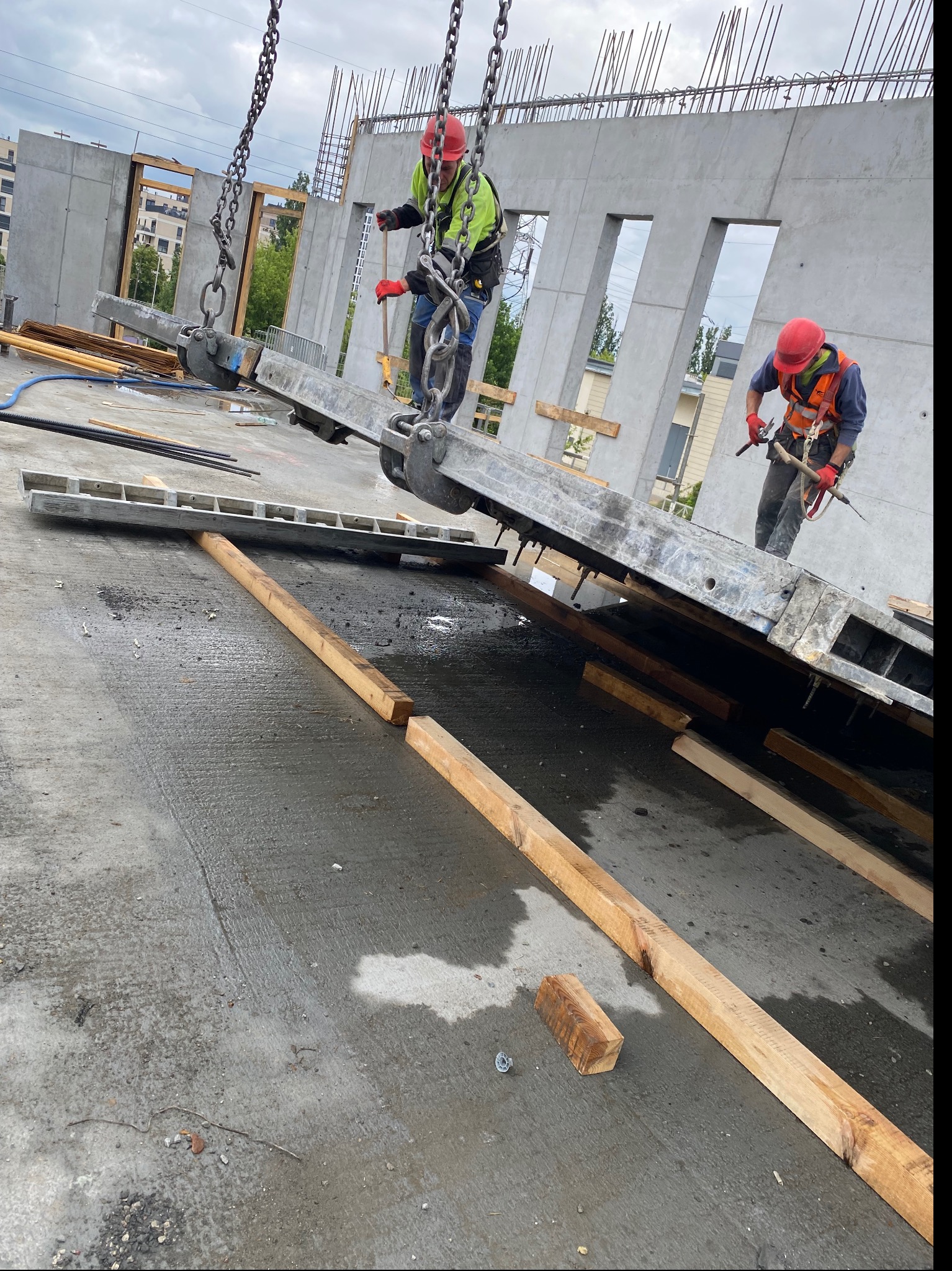 Montaż prefabrykowanej ściany betonowej z otworami okiennymi przy użyciu dźwigu łańcuchowego, dwaj pracownicy w kaskach i kamizelkach odblaskowych przy pracy, widoczne elementy zbrojenia.