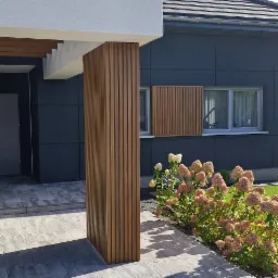 Nowoczesny dom z szarą elewacją, drewnianą kolumną z pionowych listew i panelami dekoracyjnymi, oraz ścieżką z szarych płyt chodnikowych i rabatą z kwitnącymi hortensjami.