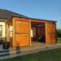 Nowoczesna, drewniana konstrukcja z przesuwnymi panelami, tworząca zadaszoną przestrzeń wypoczynkową z widocznym zestawem mebli ogrodowych w tle, na tle domu z ciemnym dachem.
