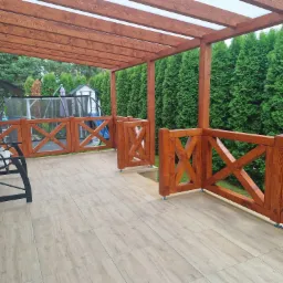 Zadaszony taras z drewnianą pergolą i balustradą, wykończony jasnymi płytkami. W tle zielone tuje i huśtawka ogrodowa.