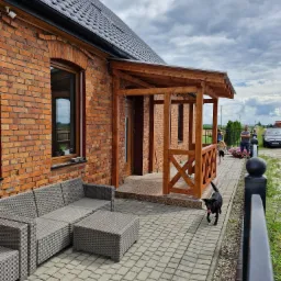 Ceglany dom z drewnianą pergolą i szarą sofą na kostce brukowej. W tle widoczni ludzie i samochód. Czarny pies biegnie w stronę widza.