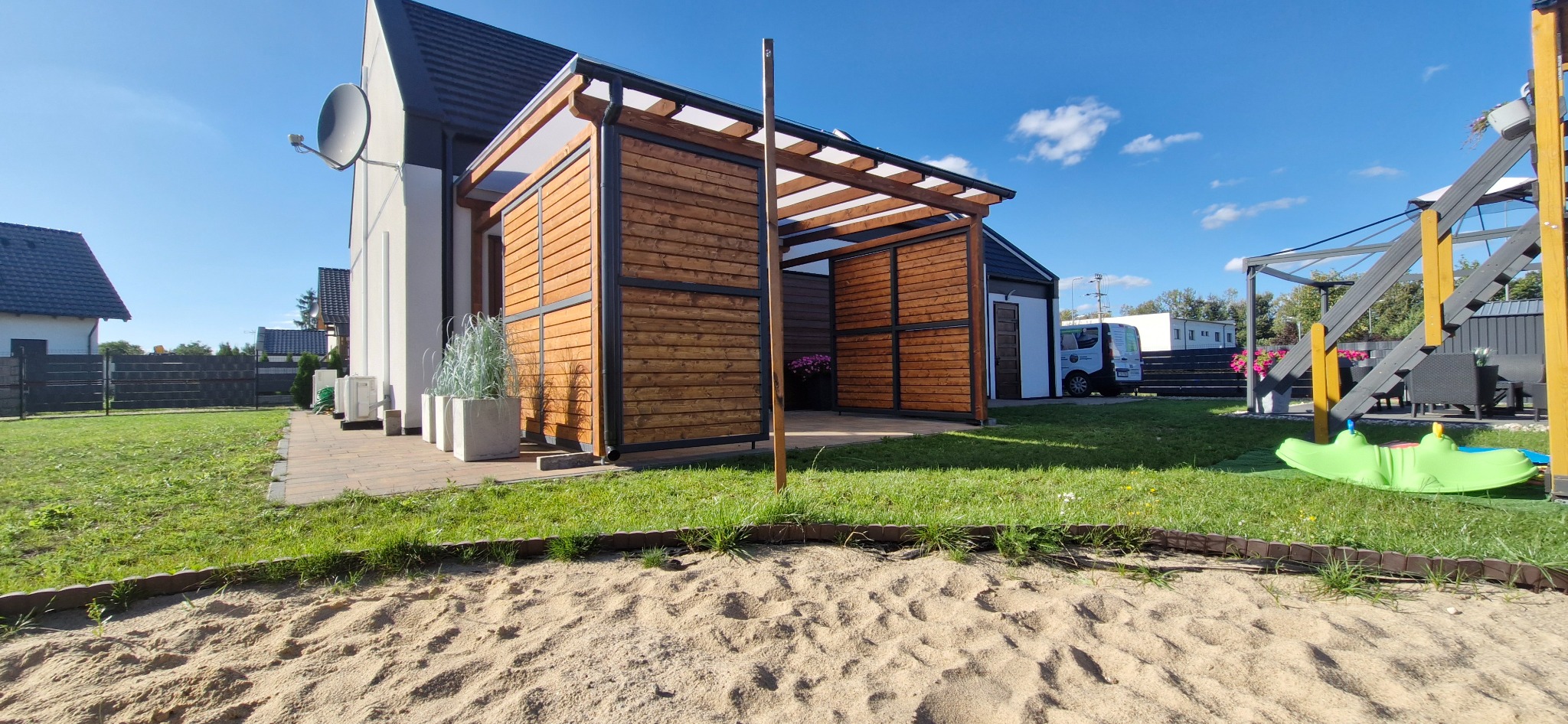 Nowoczesna pergola z drewnianymi panelami i czarną ramą, przylegająca do domu, z widokiem na ogród i piaskownicę w pierwszym planie.
