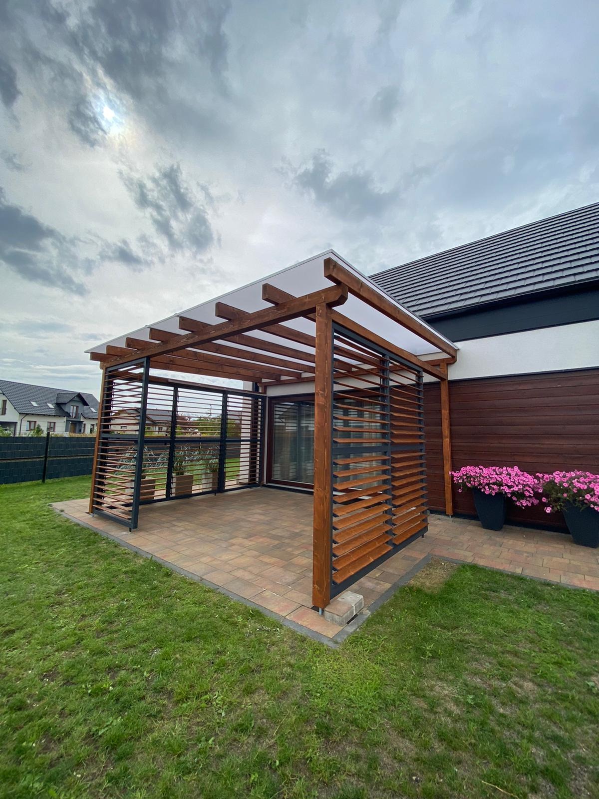 Nowoczesna pergola z drewnianymi lamelami i przezroczystym zadaszeniem, przylegająca do domu z brązową elewacją i kwiatami w donicach, na tle zielonego trawnika i pochmurnego nieba.