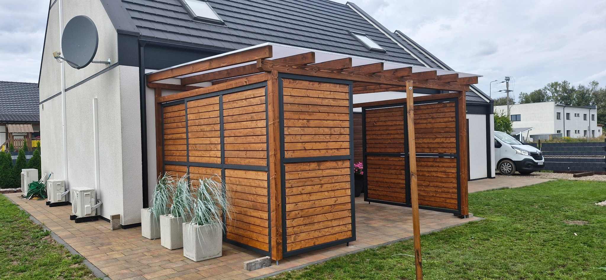 Nowoczesna pergola drewniana z poziomymi deskami w kolorze brązowym, z czarną metalową ramą, przylegająca do białego budynku z anteną satelitarną, z trzema betonowymi donicami z trawami...