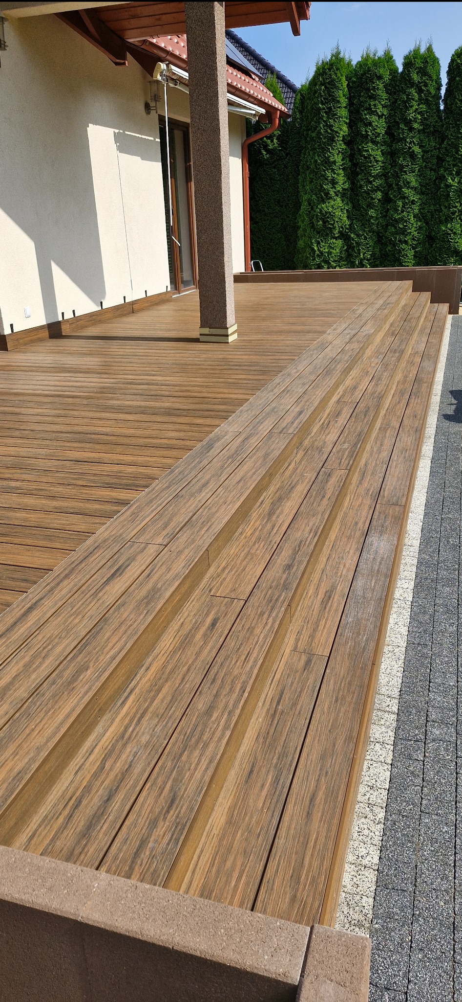 Brązowy taras kompozytowy z trzema schodkami, widok z góry, z kolumną podtrzymującą zadaszenie i obrzeżem z kostki brukowej.