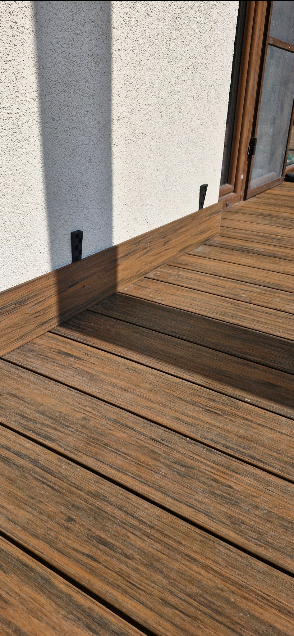 Fragment brązowego tarasu z desek, z widocznymi czarnymi elementami mocującymi przy ścianie budynku z oknem.