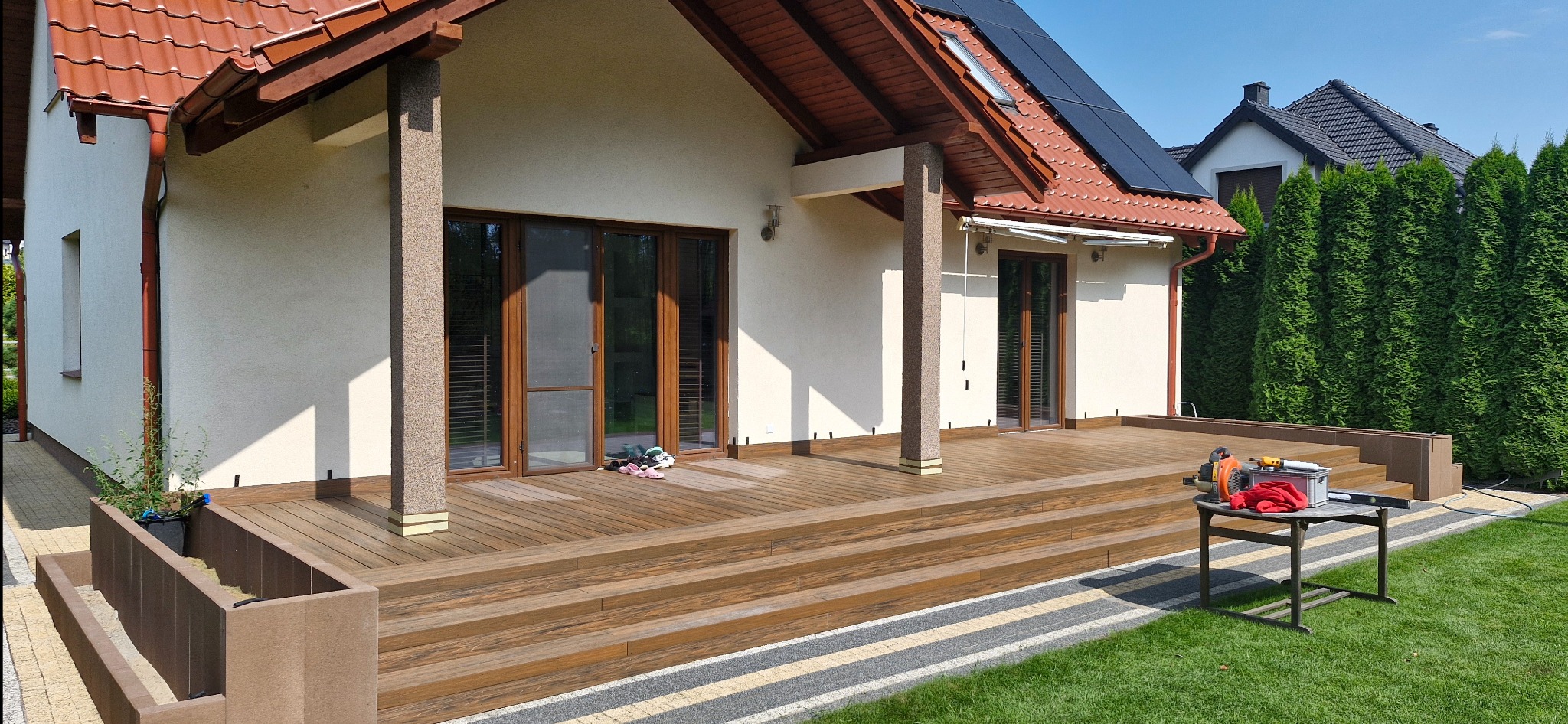 Nowoczesny dom z tarasem z desek kompozytowych, na którym widoczne są narzędzia i akcesoria do montażu, sugerujące trwające prace wykończeniowe.