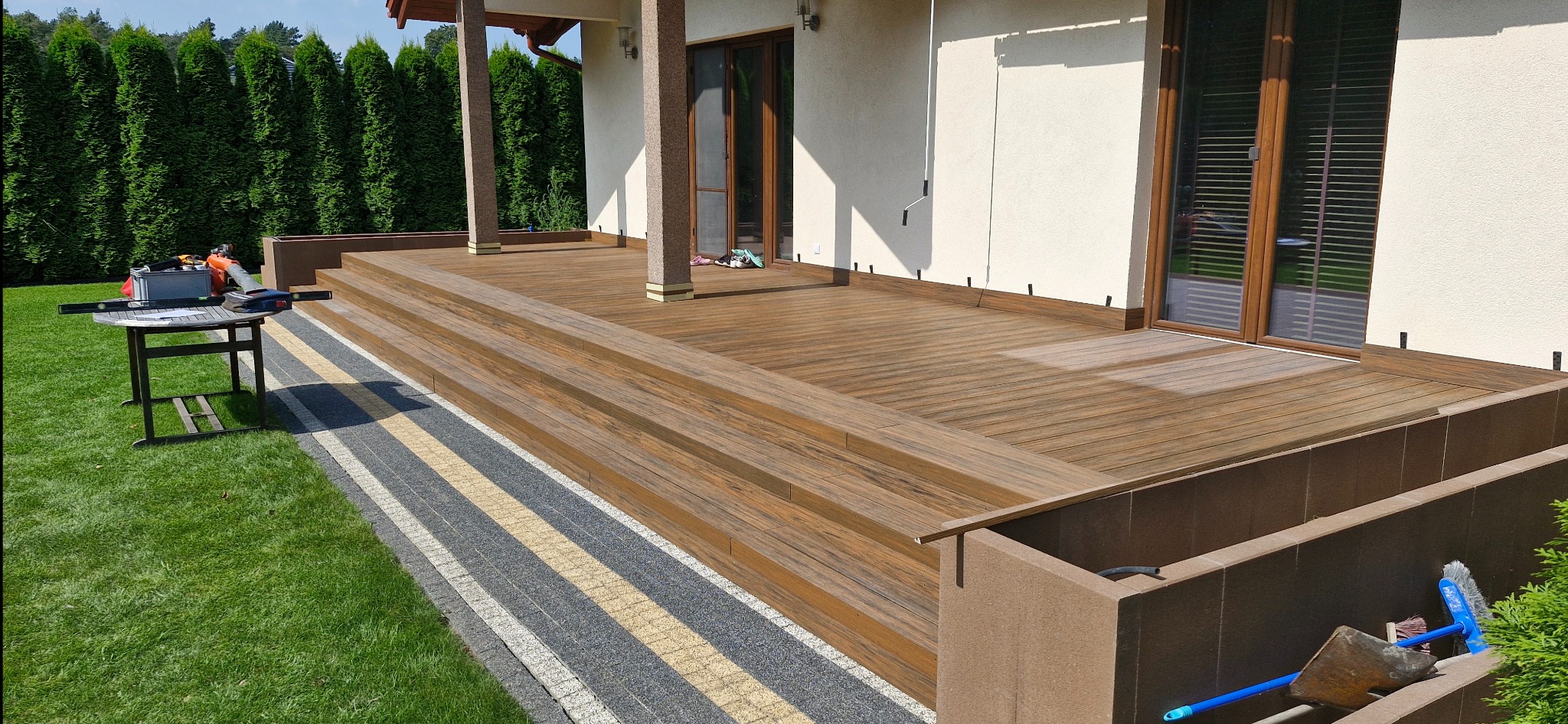 Wykończony taras z drewnianą deską, z trzema stopniami, widok na dom z brązowymi oknami, obok narzędzia na stole i kostka brukowa.