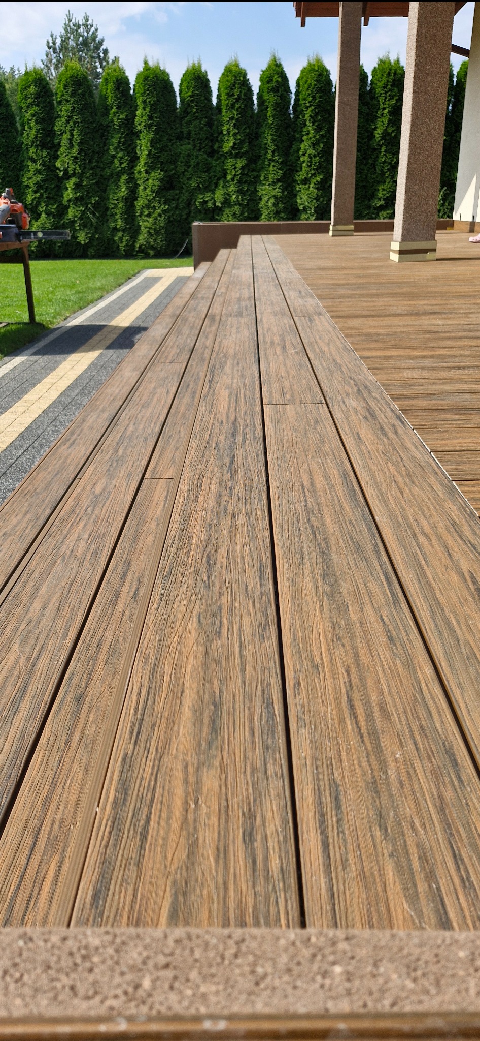 Nowoczesny taras z desek kompozytowych o fakturze drewna, z widocznym fragmentem ogrodu i tujami w tle.