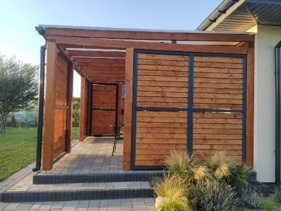 Drewniana konstrukcja z przesuwnymi panelami, tworząca zacienione miejsce do siedzenia przylegające do domu, z niskimi schodami z kostki brukowej i ozdobnymi trawami.