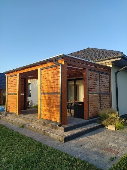 Zadaszona konstrukcja z drewnianymi panelami i metalową ramą, tworząca otwartą przestrzeń na zewnątrz domu, z widocznymi schodami wejściowymi i trawnikiem.