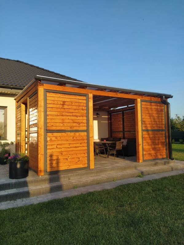 Nowoczesna, drewniana konstrukcja z przesuwnymi panelami, tworząca zadaszoną przestrzeń wypoczynkową z widocznym zestawem mebli ogrodowych w tle, na tle domu z ciemnym dachem.