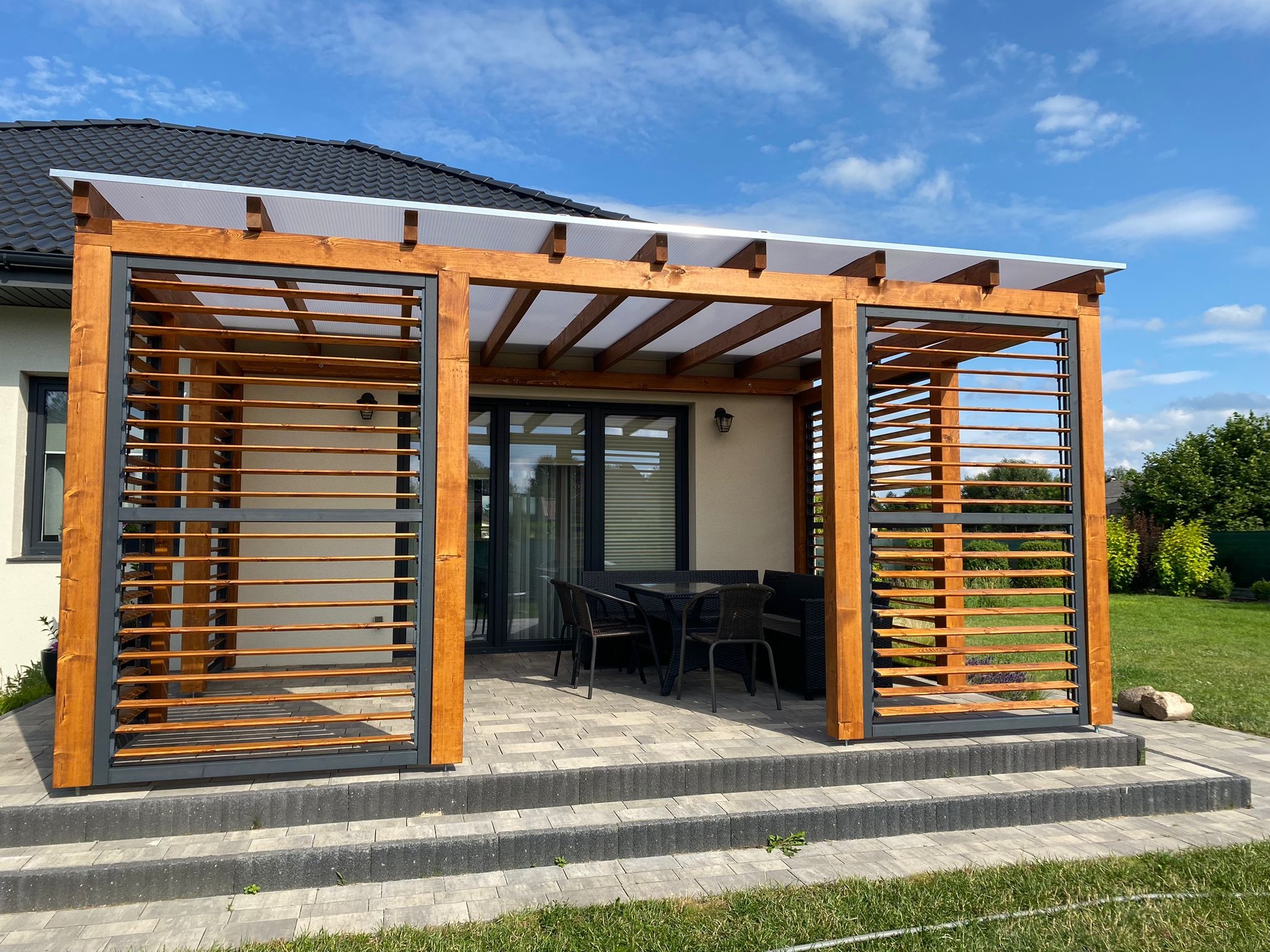 Drewniana konstrukcja z lamelami pełni rolę zadaszenia i osłony tarasu, z widocznym zestawem mebli ogrodowych i schodami z kostki brukowej.