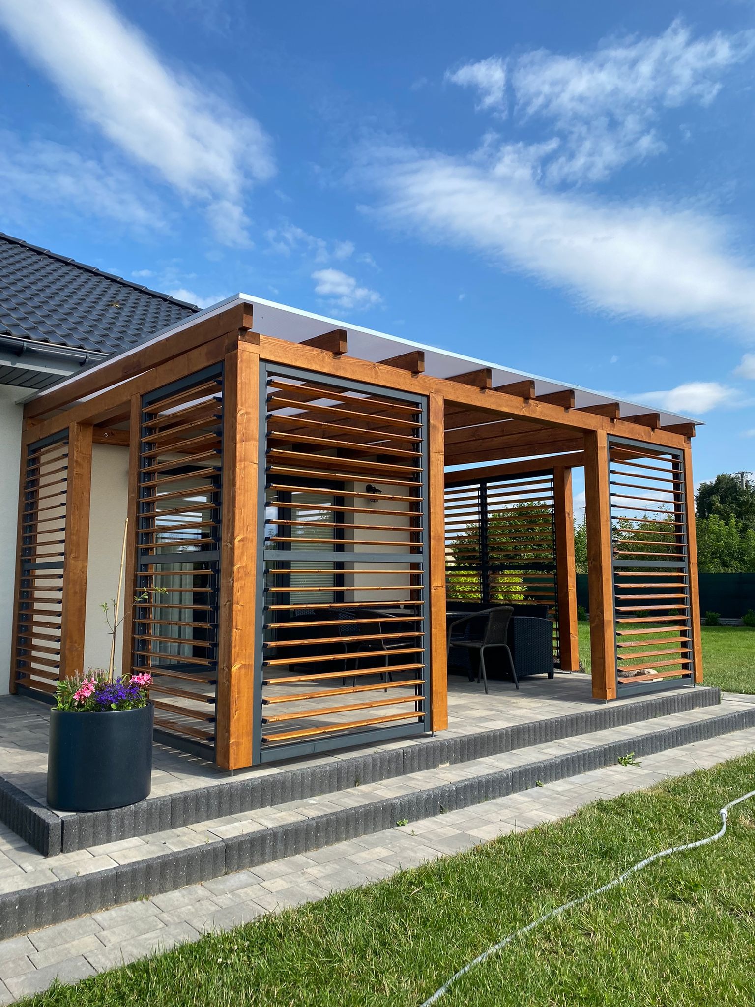Drewniana pergola z lamelami w kolorze orzecha włoskiego, zadaszona przezroczystym poliwęglanem, z widocznym kompletem mebli ogrodowych w tle, na podwyższeniu z kostki brukowej z donicą z kwiatami...
