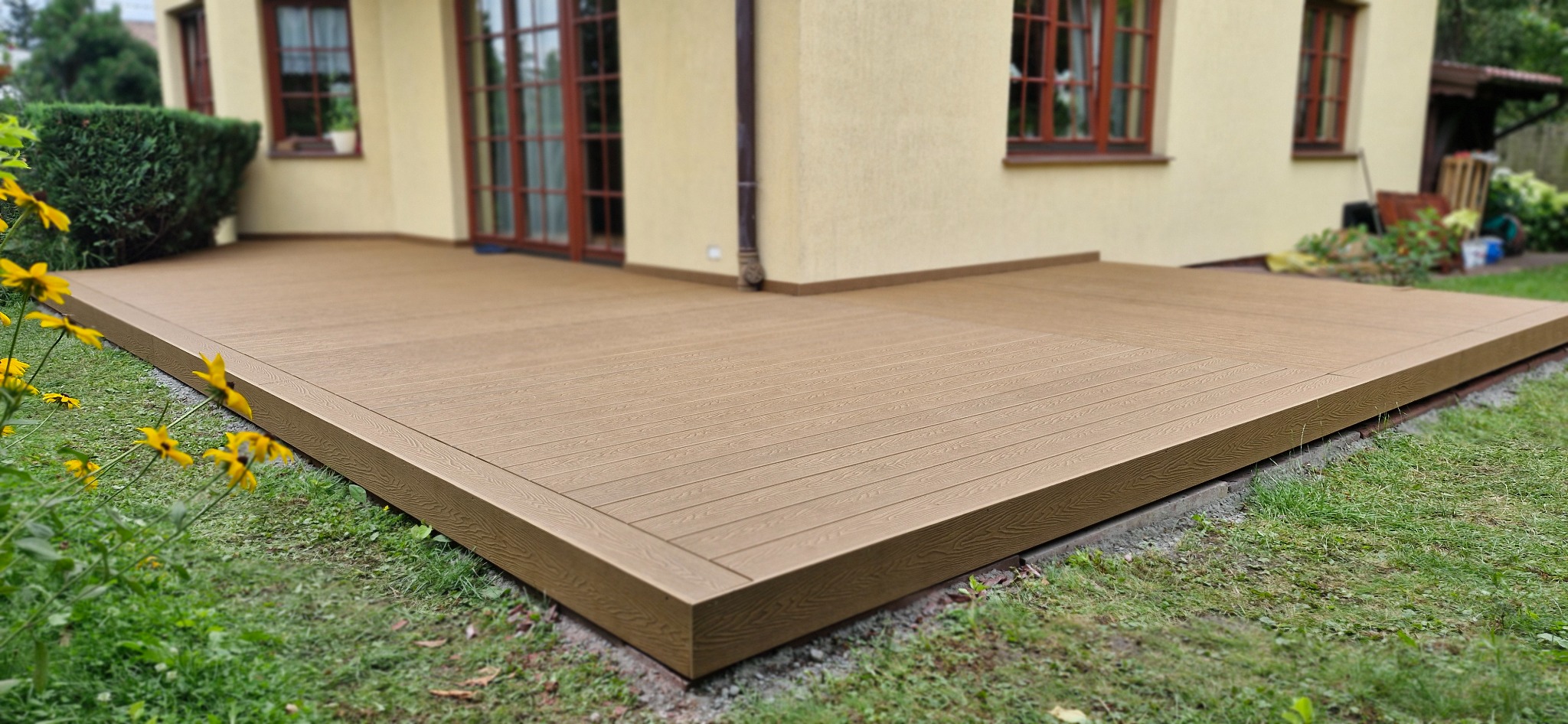 Beżowy taras z desek kompozytowych przylegający do domu, widok z poziomu trawnika z żółtymi kwiatami w pierwszym planie.