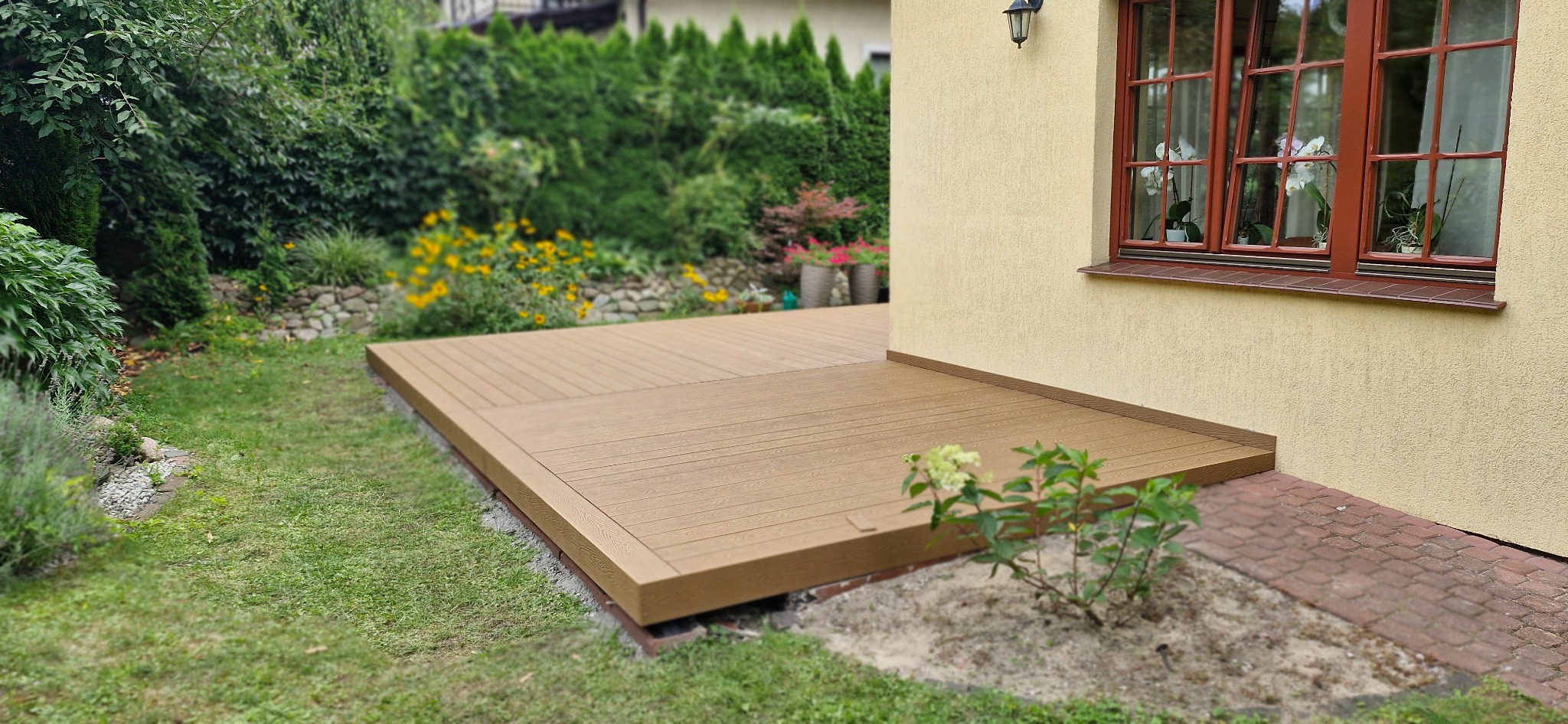 Beżowy taras z desek kompozytowych przylegający do ściany domu z oknem w brązowej ramie, otoczony zielenią ogrodu.