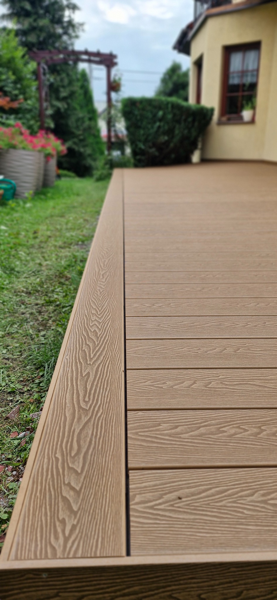 Jasnobrązowy taras z desek kompozytowych, widok z boku, zarys budynku w tle